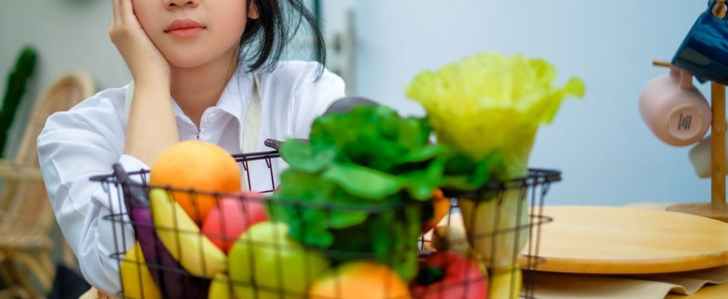 a girl sitting at a table with a basket of fruit and vegetables