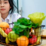 a girl sitting at a table with a basket of fruit and vegetables