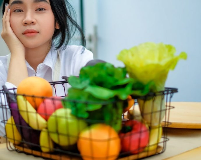 a girl sitting at a table with a basket of fruit and vegetables