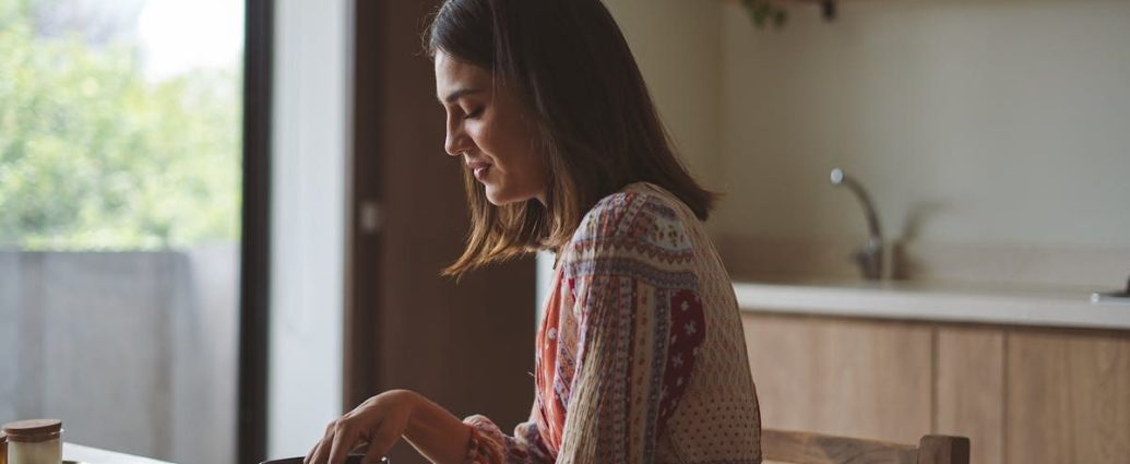 Woman in patterned dress enjoying breakfast at home near a sunlit window.