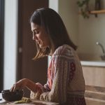 Woman in patterned dress enjoying breakfast at home near a sunlit window.